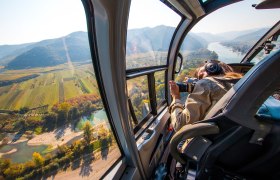 Ausblick aus dem Helikopter, © Helikopter Tours Austria Blick aus einem Helikopter auf eine Landschaft mit Fluss und Feldern.
