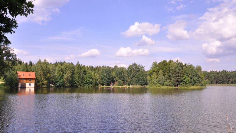 Mitterteich, © Marktgemeinde Hohneich Ein See mit einem Haus am Ufer, umgeben von Wald und blauem Himmel mit Wolken.