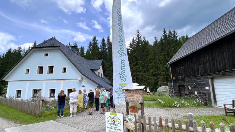 Kräutergarten Kanlehen, © "Natur im Garten" Besuchergruppe vor einem blauen Haus im Kräutergarten Kanlehen mit Fahne und Schildern.
