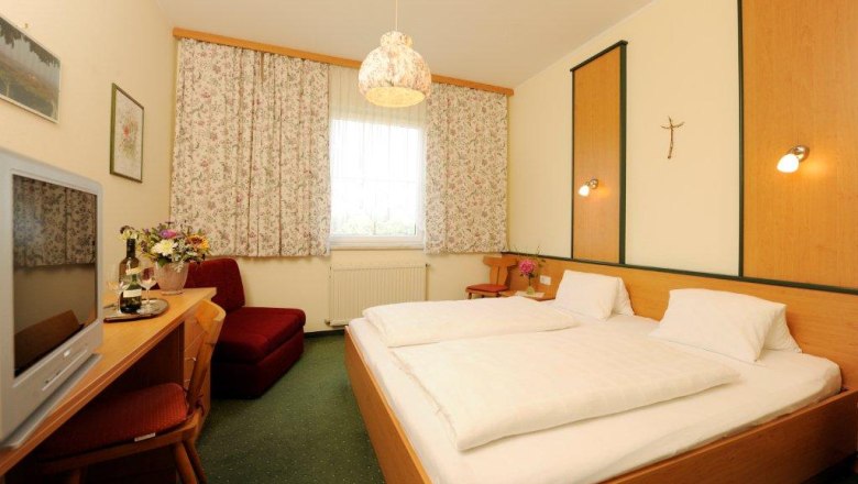 Zimmerbeispiel, © Fam. Rethaller Gemütliches Hotelzimmer mit Doppelbett, TV, Blumen und Vorhängen.