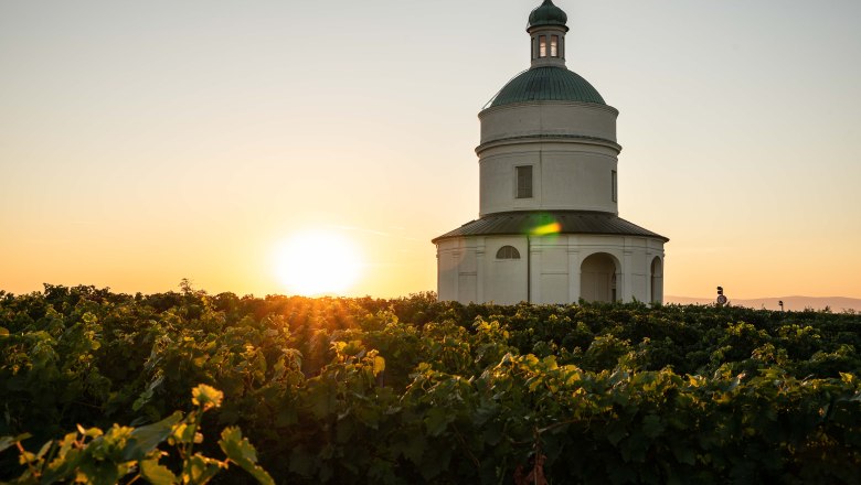 Mannersdorf, © www.paulbreuss.com Eine weiße Kapelle steht inmitten eines Weinbergs bei Sonnenuntergang.