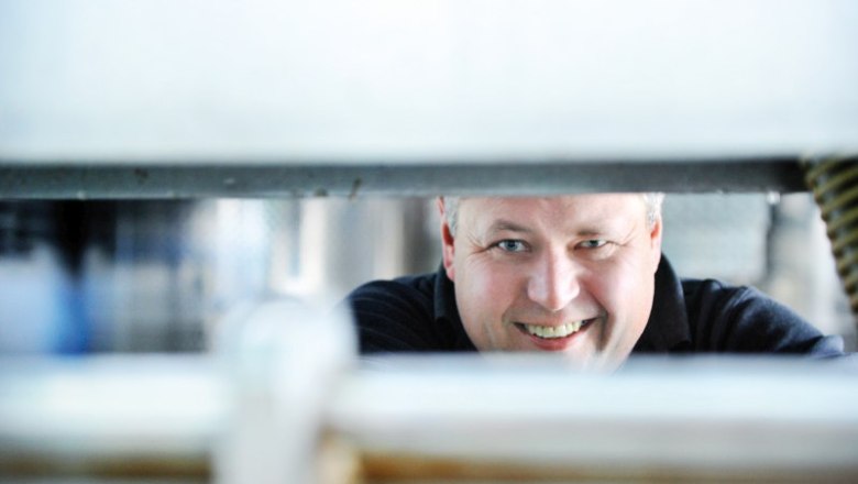 weingut-jschmid, © Weingut Josef Schmid / pov.at Ein Mann lächelt durch einen schmalen Spalt in einer industriellen Umgebung.