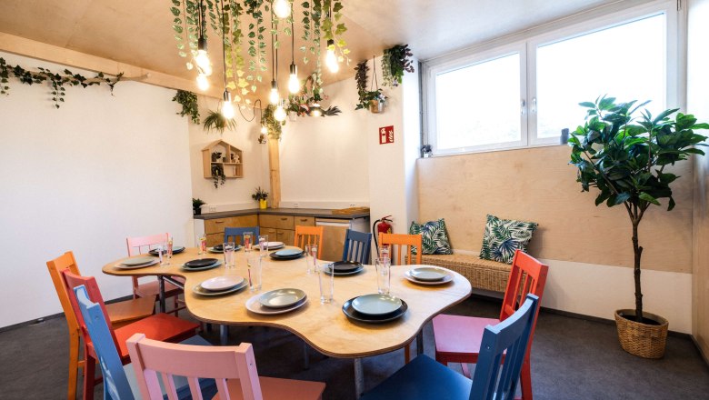 BigWall_partyyyyy, © bigwall Ein gemütlicher Raum mit einem Tisch, umgeben von bunten Stühlen und Pflanzen.