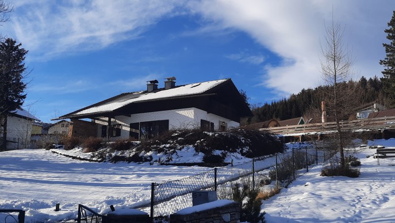 Belle Air Lodge im Winter, © Christoph Gierlinger Ein verschneites Haus mit dunklem Dach und Schornsteinen, umgeben von Bäumen und blauem Himmel.
