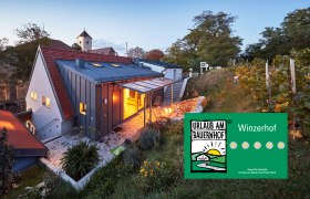 Weinviertler Kellerstöckl, © Andreas Hofer Ein modernes Kellerstöckl im Weinviertel bei Dämmerung, umgeben von Weinreben und einem Garten. Ein Schild zeigt 'Urlaub am Bauernhof'.