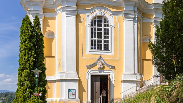 Bergkirche Pitten, © Wiener Alpen, Christian Kremsl Gelbe Barockkirche mit weißen Säulen und einer Statue über dem Eingang, umgeben von Bäumen und blauem Himmel.
