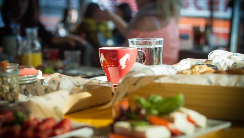 2Stein Bar-Café-Restaurant, © 2stein_Kastner new media Ein roter Kaffeebecher und ein Glas Wasser auf einem Tisch in einem Café, umgeben von Essen und Getränken.