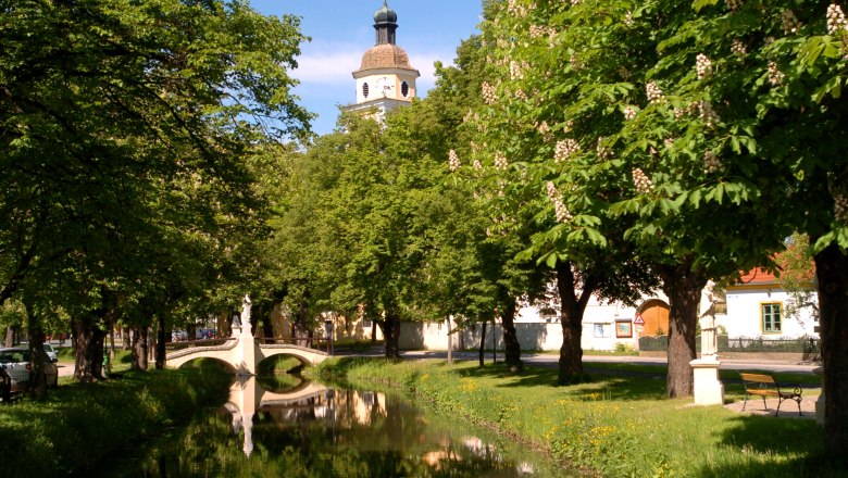 Gemeinde Straß im Straßertale, © Gemeinde Straß im Straßertale Ein idyllischer Park mit einem kleinen Fluss, einer Brücke und einem Kirchturm im Hintergrund.