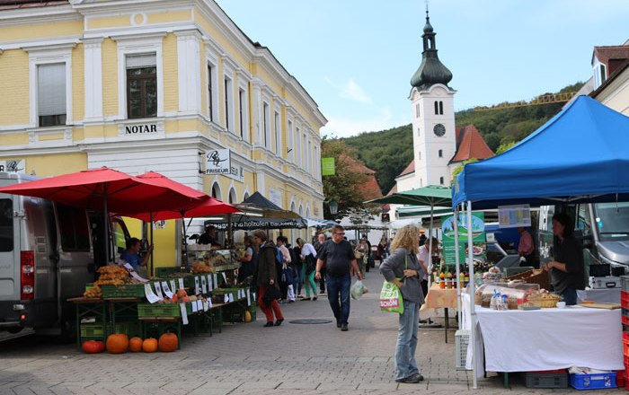 Bauernmarkt, © Wienerwald Tourismus GmbH Ein belebter Bauernmarkt mit Ständen und Besuchern vor einem gelben Gebäude und einer Kirche im Hintergrund.