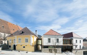 Mauritiushof Weinerlebniswelt, © Weingut FJ Gritsch Zwei Gebäude mit gelber und weißer Fassade, verbunden durch einen modernen Glasgang, unter blauem Himmel.