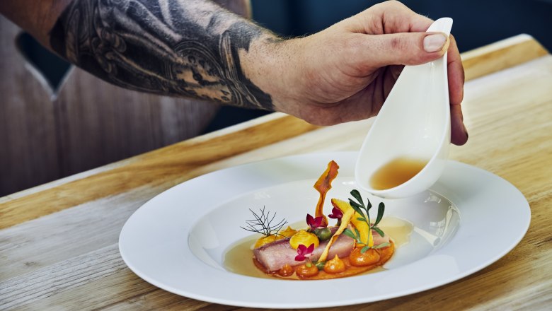 Hier isst das Auge mit, © Niederösterreich Werbung/Andreas Hofer Ein Teller mit kunstvoll angerichtetem Essen, über das eine Hand aus einer kleinen Kanne Sauce gießt.