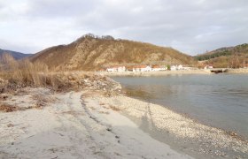 strand, © Kowalski Ein ruhiger Strand mit Kieselsteinen, im Hintergrund ein Dorf und bewaldete Hügel.