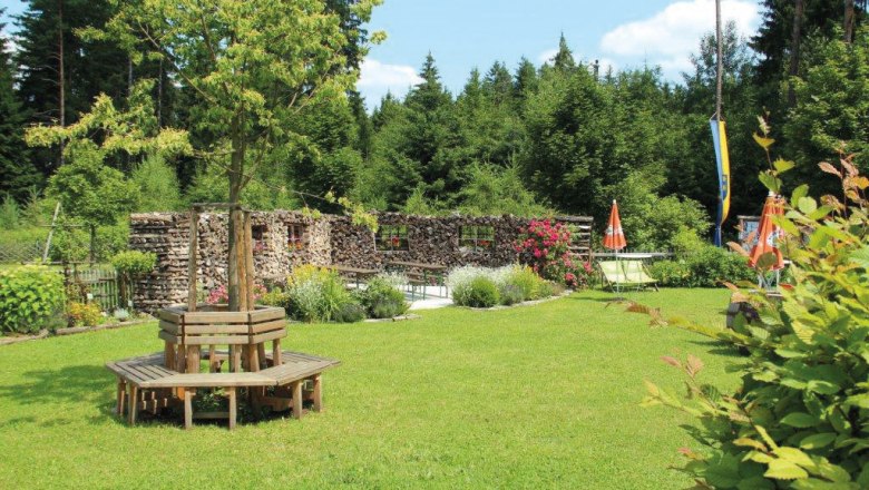 Waldschenke Schreiber, © Waldschenke Schreiber Ein grüner Garten mit Holzbänken um einen Baum, umgeben von Wald und Blumen.