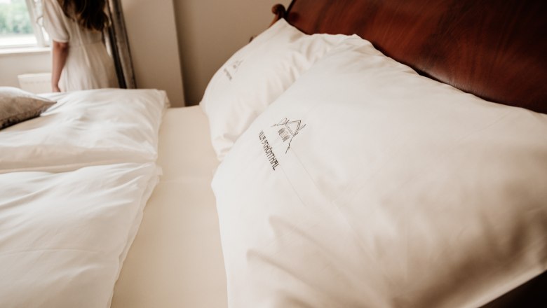 villa_schonthal-20_2, © Julius Hirtzberger Nahaufnahme eines Bettes mit weißen Kissen und Bettwäsche, im Hintergrund eine Frau am Fenster.