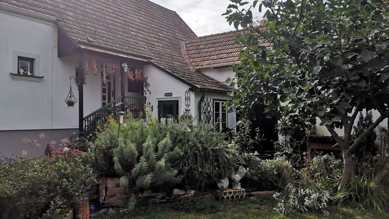 Scherben, Blüten, Blatt und Feder, © "Natur im Garten" Ein gemütliches Haus mit Garten, umgeben von Pflanzen und einem Baum.