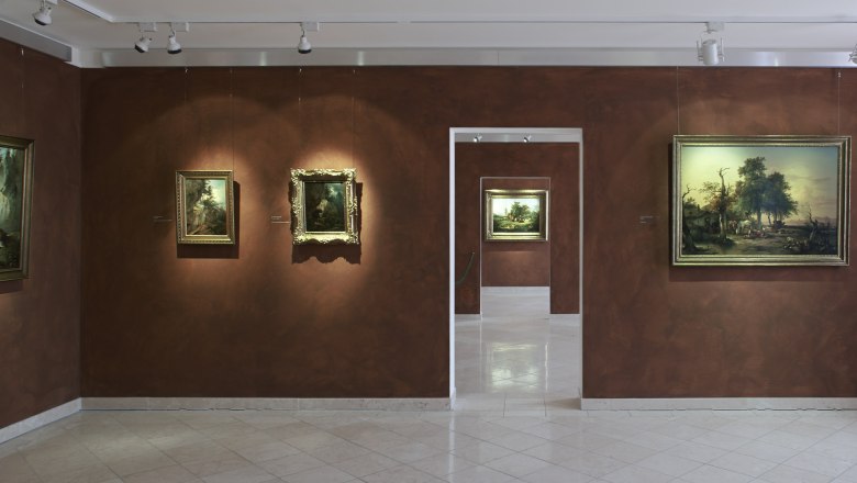 Ausstellungsräume im Gauermann Museum, © Wiener Alpen, Foto: Bene Croy Innenansicht eines Museums mit Gemälden an braunen Wänden.