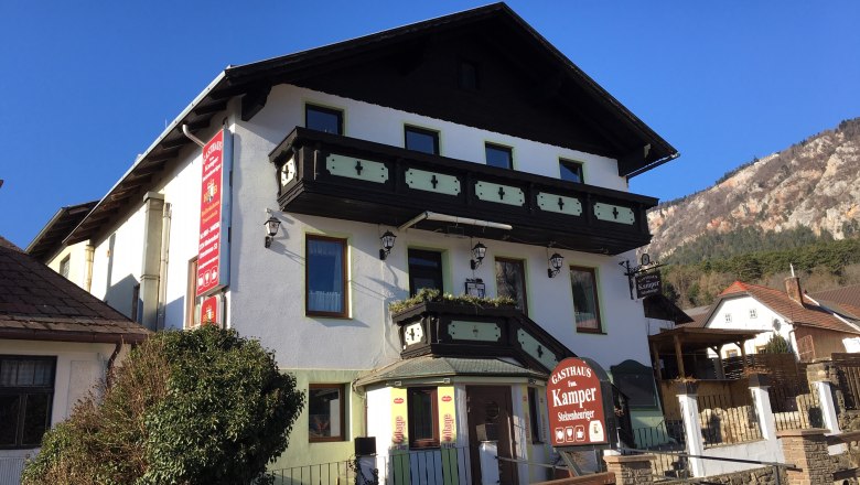 Stelzenheuriger Familie Kamper, © Familie Kamper Ein traditionelles Gasthaus mit Holzverzierungen und einem Schild "Gasthaus Fam. Kamper" vor einem klaren blauen Himmel.