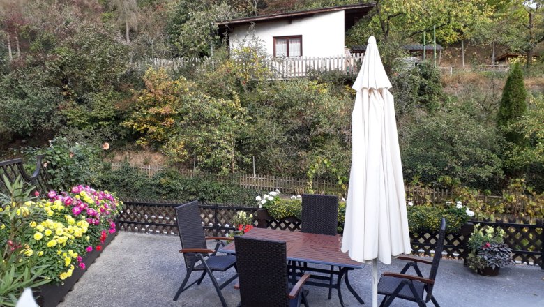 Terrasse bei Familie Lindenhofer, © Familie Lindenhofer Terrasse mit Tisch, Stühlen und Sonnenschirm, umgeben von Blumen und Bäumen.