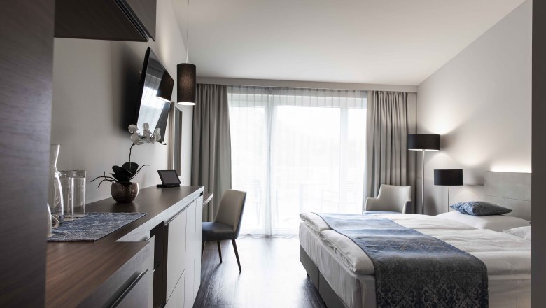 Zimmer, © NMC Modernes Hotelzimmer mit Doppelbett, Fernseher, Schreibtisch und großen Fenstern mit Vorhängen.