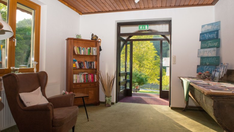 Eingangsbereich / Lobby, © Dorfschmiede eG Eingangsbereich mit Sessel, Bücherregal und offenem Türbereich mit Blick ins Grüne.