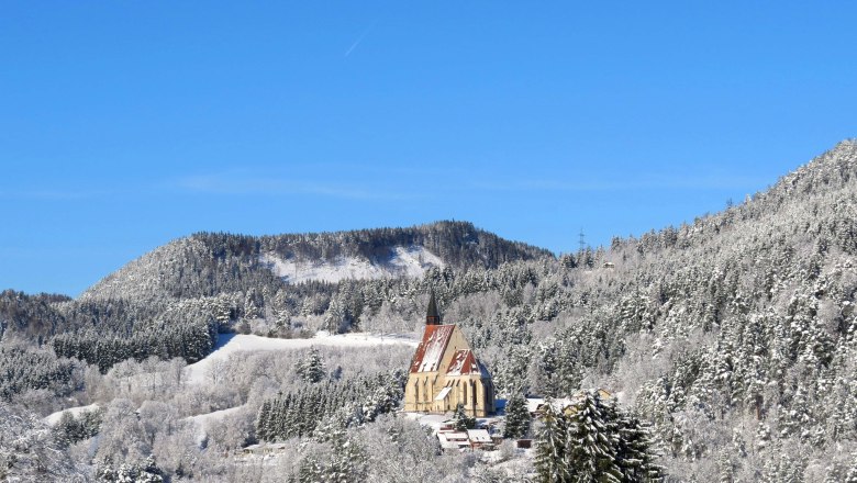 Die Wolfgangskirche thront über dem Ort, © Marktgemeinde Kirchberg, Wolfgang Riegler Winterlandschaft mit Kirche in verschneiten Bergen unter blauem Himmel.