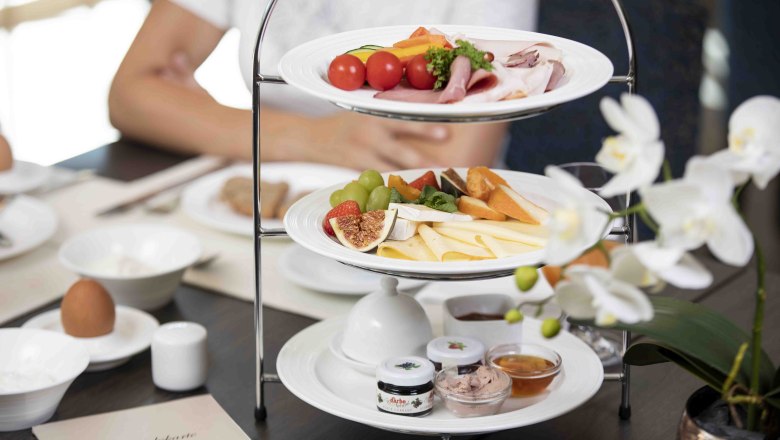 Frühstück, © NMC Ein Frühstückstisch mit einer Etagere voller Käse, Aufschnitt, Obst und Brotaufstrichen. Im Vordergrund eine weiße Orchidee.