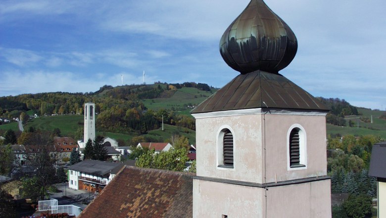 Marktgemeinde Traisen, © zVg Marktgemeinde Traisen Kirchturm mit Zwiebeldach vor hügeliger Landschaft und Windrädern.
