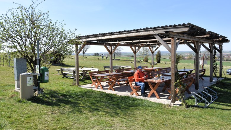 Genussrastplatz beim Baumkreis, © StadtGemeinde Mistelbach / Mag. Mark Schönmann Ein überdachter Rastplatz mit Holzbänken und Tischen auf einer Wiese, eine Person sitzt dort. Im Hintergrund sind Felder und Bäume zu sehen.