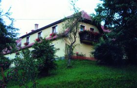 Liebhaber, © Liebhaber Ein gelbes Haus mit roten Dachziegeln und Blumenkästen an den Fenstern, umgeben von Bäumen und einem grünen Hang.