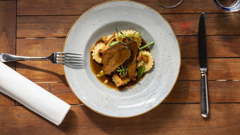 Geschmorte Rindsbackerl, © Niederösterreich Werbung/Michael Reidinger Ein Teller mit geschmorten Rindsbackerl, Ravioli und Gemüse auf einem Holztisch, daneben Besteck und ein Glas Rotwein.