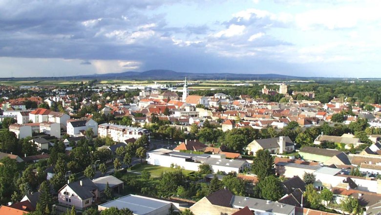 Bruck an der Leitha, Römerland Carnuntum, © Wolfgang Stinauer Luftaufnahme von Bruck an der Leitha mit Häusern und Kirche, umgeben von grüner Landschaft und bewölktem Himmel.