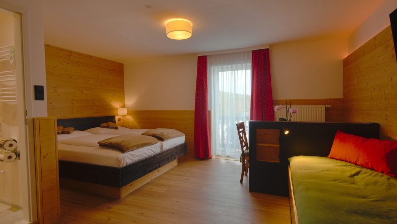 Doppelzimmer, © Alpengasthof Enzian Ein gemütliches Doppelzimmer mit Holzwänden, einem Doppelbett, einem Sofa und einem Schreibtisch. Ein Fenster mit roten Vorhängen lässt Tageslicht herein.