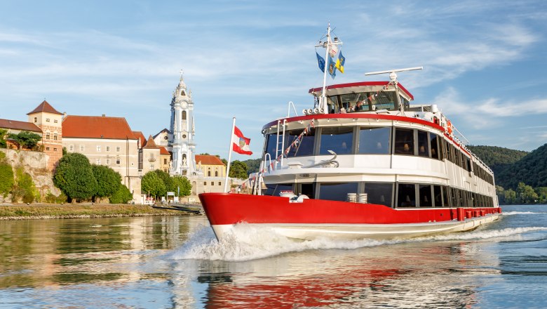 DDSG Blue Danube Schiffahrt, © DDSG Blue Danube Schiffahrt/Wöckinger Ein rotes Passagierschiff fährt auf der Donau vor einer historischen Stadt mit einem markanten Kirchturm.