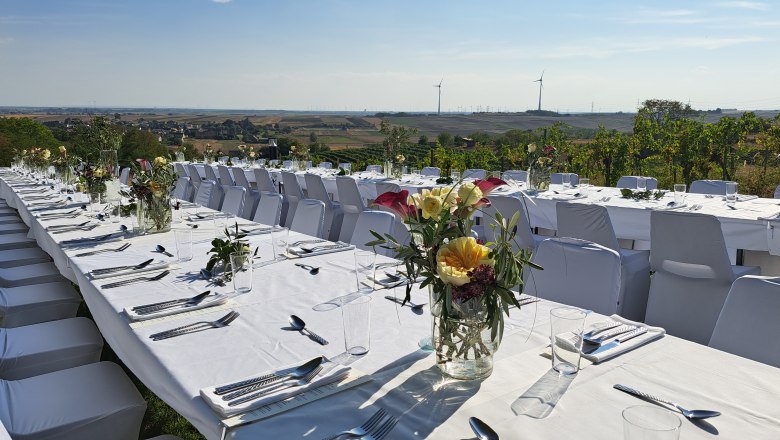 Tafeln im Weinviertel, © Weinviertel Tourismus GmbH Lange, festlich gedeckte Tische im Freien mit Blumenarrangements und Blick auf eine Landschaft mit Windrädern.