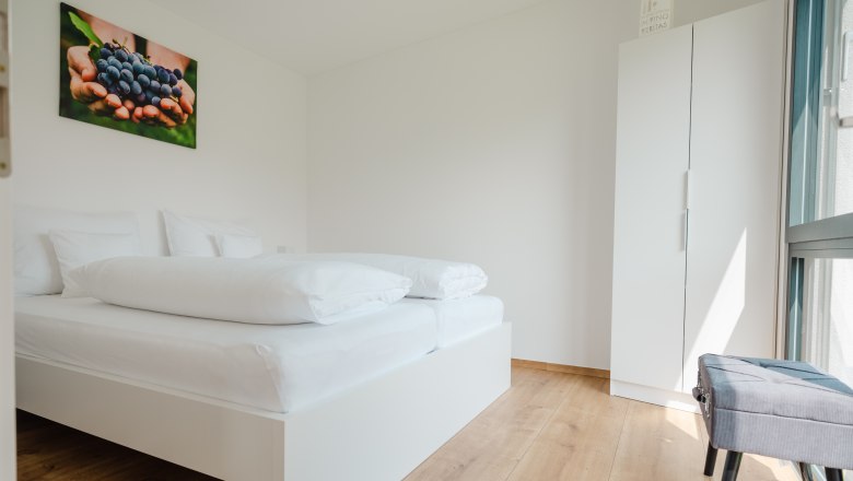 Ipsmiller Appartements, © Michael Reidinger Modernes Schlafzimmer mit weißem Bett, Holzfußboden und Bild von Trauben an der Wand.
