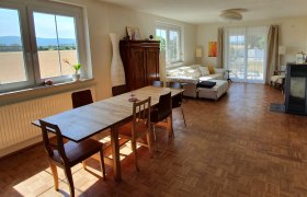 Zimmeransicht, © Ferienhaus Kerschbaum Helles Zimmer mit Esstisch, Stühlen, Sofa und großen Fenstern mit Blick auf die Landschaft.