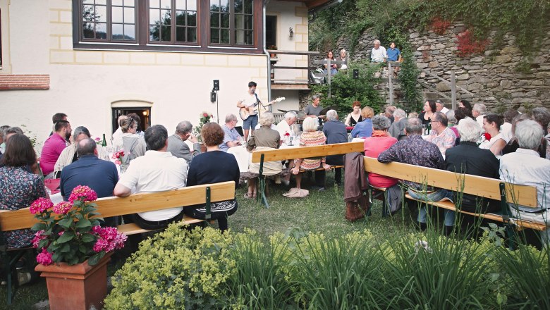 Gastgarten Heuriger, © Monika Pölzer Menschen sitzen im Freien an langen Tischen bei einem Heurigen, während ein Musiker auftritt.