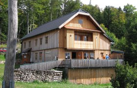Waldburgangerhütte, © Maria Stroebl Eine Holzhütte im Wald mit Terrasse und Personen darauf.