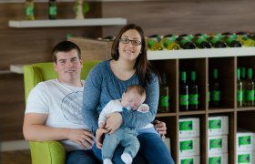 Familie Lust, © Astrid Bartl Ein Paar mit einem Baby sitzt auf einem grünen Sessel in einem Weingut.