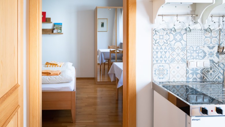 Weingut Mehofer - Neudeggerhof, © Elisabeth Mandl Innenansicht eines Zimmers mit Bett, Spiegel, Tisch und kleiner Küche mit gemusterten Fliesen.