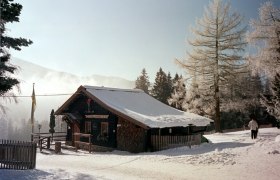 Grenzlandhütte, © Grenzlandhütte, Foto Paul Laschitz Eine verschneite Berghütte in winterlicher Landschaft mit Bäumen und einem Spaziergänger.