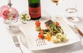 Wachauerstube, © Rita Newman Elegantes Gericht mit Fisch, Spargel und Kartoffeln auf weißem Teller, dekoriert mit Blumen. Im Hintergrund eine Flasche Wein und ein Glas.