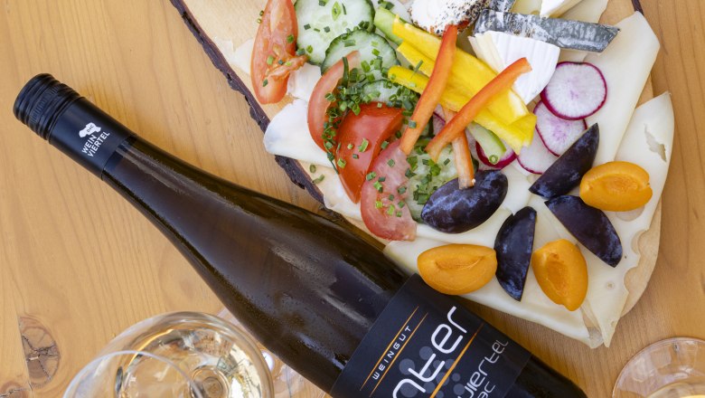 Heurigenplatte, © Weinviertel Tourismus GmbH / Schwarz-König Eine Heurigenplatte mit verschiedenen Käsesorten, Aufstrichen, Gemüse und einer Flasche Wein auf einem Holztisch.