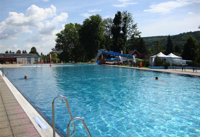 Schlossbad Pitten, © Marktgemeinde Pitten Ein großes Freibad mit klarem Wasser, umgeben von Bäumen und Hügeln unter einem blauen Himmel.