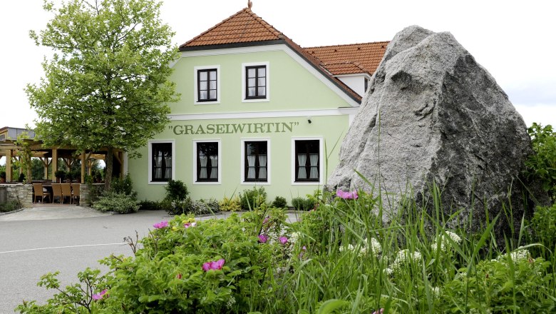 Graselwirtin, © Gasthof Graselwirtin Grünes Gebäude mit der Aufschrift 'Graselwirtin', umgeben von Pflanzen und einem großen Felsen im Vordergrund.