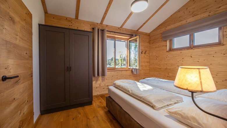 Kittenberger Chalets am Gartensee, © Niederösterreich Werbung / Maximilian Pawlikowsky Innenansicht eines Schlafzimmers in einem Chalet mit Holzwänden, einem Doppelbett, einem Kleiderschrank und einem Fenster mit Blick nach draußen.
