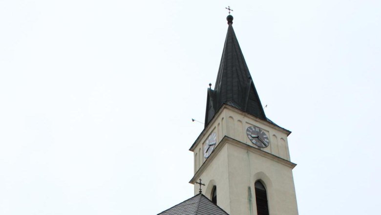 Guntersdorf, © Gemeinde Guntersdorf Kirchturm mit Uhr und spitzem Dach vor hellem Himmel.