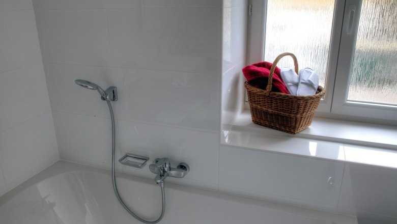Badewanne im Loft 1, © Carlos de Mello Weiße Badewanne mit Duschkopf, daneben ein Fensterbrett mit einem Korb voller Handtücher.