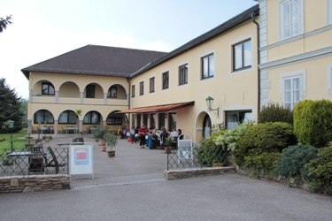 Naglhof, © Naglhof Ein gelbes Gebäude mit Arkaden und Terrasse, auf der Menschen sitzen.