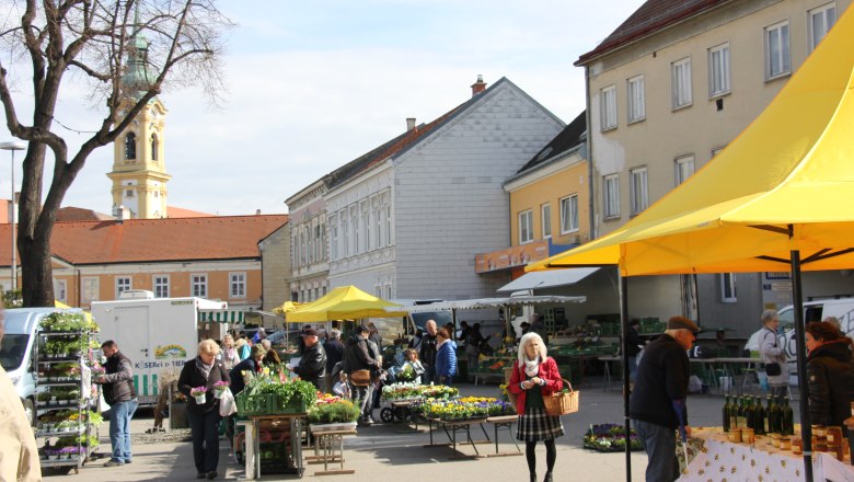 Wochenmarkt Stockerau, © Johannes Ehn Menschen auf einem Wochenmarkt in Stockerau mit gelben Marktständen und einer Kirche im Hintergrund.